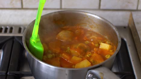 Close-up-of-boiled-greek-tomato-pea-stew-with-artichokes-and-potatoes-,-man-stirring-with-a-silicone-spoon,-slow-motion