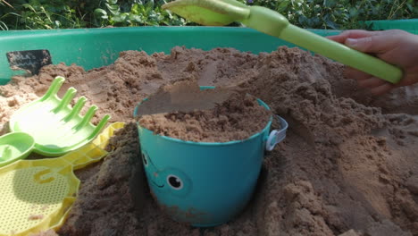 Child-patting-a-bucket-full-of-sand-with-a-spade,-shot-in-a-children's-sand-pit