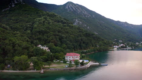 A-luxurious-villa-with-a-swimming-pool-and-a-stone-pier-on-the-shores-of-Kotor-bay-in-Montenegro,-surrounded-by-trees-and-a-steep-mountainside,light-evening-glow-over-the-area-and-the-bay-waters