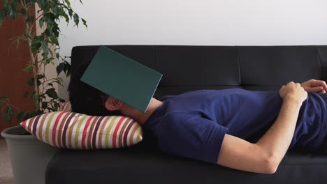 Man-lying-on-the-couch-sleeping,-with-his-face-covered-by-a-book