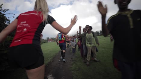 Marathon-Athleten-Laufen-Hintereinander-Auf-Einer-Unbefestigten-Straße,-Geben-Den-Kindern-Unterwegs-High-Fives-Und-Lächeln-In-Den-Buschdschungelfeldern-Tansanias-–-Zeitlupe