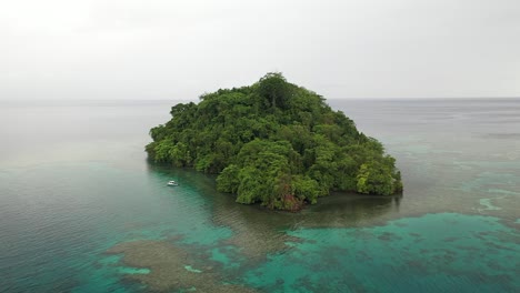 Drone-shot-backwards-of-a-small,-lush-and-green-island-with-a-small,-white-boat-next-to-it