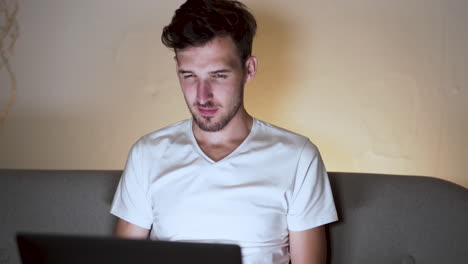 A-young-man-with-a-stubble-and-a-modern-hairstyle,-wearing-a-white-t-shirt,-sitting-at-home-on-a-sofa,-chatting-with-someone-on-a-laptop,-smiling-and-laughing,-amused-by-the-reply,-static-close-up-4k