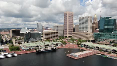 Descending-aerial-of-Baltimore-Inner-Harbor-during-sunny-summer-day,-tall-skyscrapers-and-puffy-clouds-fill-sky-at-waterfront-tourist-attraction