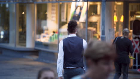 An-elegant-young-guy-wearing-a-simple-green-protective-Covid-19-facemask,-a-black-waistcoat,-white-shirt-and-jeans-crossing-the-street,-his-back-to-the-camera,-high-end-shop-in-the-background,-4k