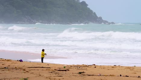 Phuket,-Thailand---September-20,-2020:-A-man-in-a-yellow-shirt-stands-fishing-on-the-beach-on-windy-days-including-the-stormy-season-in-Thailand