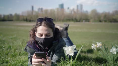Woman-wearing-a-black-face-mask-lying-on-the-grasses-and-playing-with-her-phone-in-the-park-during-a-summer-day-with-skyscrapers-in-the-horizon