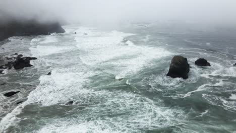 White-foamy-emerald-green-waves-crash-on-a-rocky-jagged-fog-shrouded-coastline,-aerial