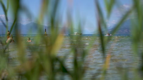 Wide-shot-of-people-swimming-in-the-lake-with-boats-passing-by,-shot-through-reeds-on-Jamaica-Beach,-Sirmione,-Lago-Garda,-Lake-Garda,-Italy