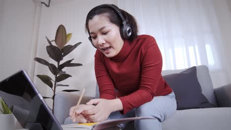 Asiatische-Frau-Auf-Rotem-Hemd,-Die-Ein-Tablet-Mit-Kopfhörer-Für-Ein-Online-Meeting-Zu-Hause-Nutzt