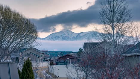Suburban-neighborhood-with-clouds-moving-in-two-layers-in-opposite-directions-in-front-of-the-snowy-mountain---zooming-time-lapse