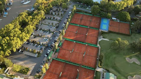 Aerial-orbiting-over-clay-tennis-courts,-a-golf-playground-and-a-parking-lot-on-a-private-sport-club-at-golden-hour