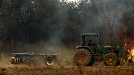 Tractor-drives-across-a-field-with-a-wildefire-burning-brush-in-the-background