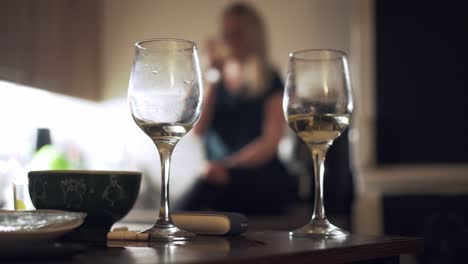 Zwei-Gläser-Weißwein-Auf-Dem-Tisch-Und-Trinkendes-Blondes-Mädchen-Im-Hintergrund,-Hausparty-Konzept