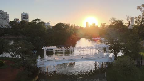Aerial-rising-over-boats-sailing-under-white-bridge-in-Rosedal-gardens,-Palermo-neighborhood-at-sunset,-Buenos-Aires