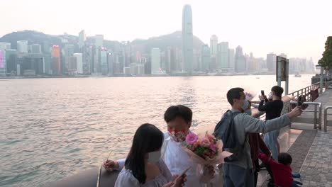 Ein-Paar-Macht-Ein-Selfie-Mit-Einer-Rosenboutique,-Während-Die-Menschen-Den-Blick-Auf-Die-Skyline-Von-Hongkong-Am-Wasser-Des-Victoria-Harbour-Genießen,-Während-Der-Sonnenuntergang-Untergeht