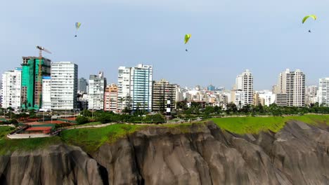 4k-aerial-video-of-3-paragliders-flying-over-the-coastline-of-the-pacific-ocean-in-the-green-area-of-Miraflores,-Lima,-Peru