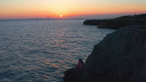 Luftaufnahme-Von-Zwei-Dominikanischen-Grüßenden-Fischern,-Die-Auf-Einer-Klippe-Sitzen,-Mit-Sonnenuntergang-Im-Hintergrund,-Pedernales