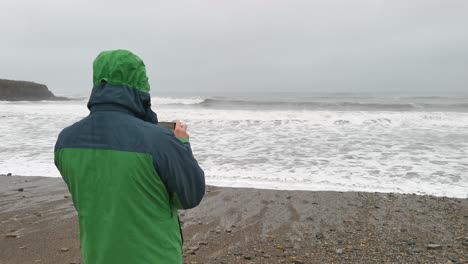 Man-standing-on-the-beach-in-stormy-weather-taking-pictures-with-waves-crashing-on-shore