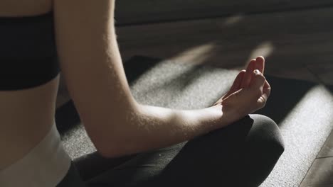 Relaxed-young-woman-in-sportswear-is-meditating-in-lotus-position-sitting-on-yoga-mat-at-home-enjoying-indoor-meditation-with-hands-on-her-knees-and-sunbeams-shining-through-the-windows