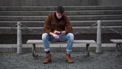Young-man-sitting-on-a-city-bench,checking-his-watch,waiting