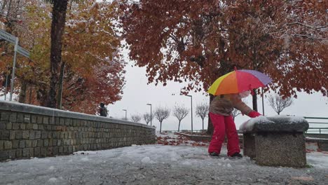 Child-girl-with-colored-umbrella-and-red-trousers-plays-kicking-snow