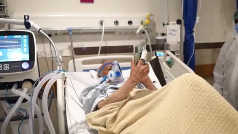Elderly-Covid-ICU-Patient-Waving-Holding-Up-Mobile-Phone-In-Hospital