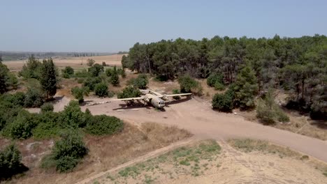 aerial-slow-forward,-zoom-in-drone-shot-of-an-old-dismantled-army-plane-monument-placed-on-an-empty-dirt-field,-surrounded-by-trees-and-bushes