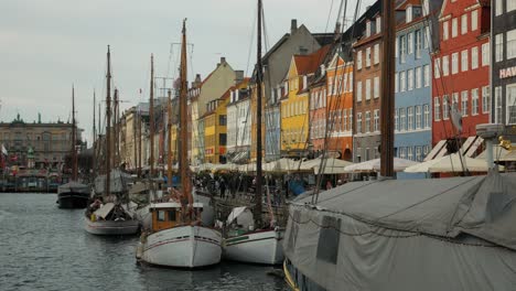 Viejos-Barcos-Amarrados-En-El-Canal-Nyhavn-Con-Gente-Caminando-Por-Casas-Adosadas-Y-Café-Con-Fachada-Colorida-En-Copenhague,-Dinamarca