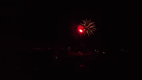 Long-slow-moving-clip-as-drone-is-slowly-flying-towards-exploding-fireworks-show-over-lake-to-see-beautiful-colors-at-night-in-the-dark