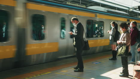 Tren-Pasando-Por-La-Plataforma-Con-Varios-Pasajeros-Con-Máscara-Protectora-En-Tokio,-Japón
