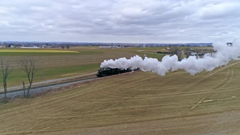 Eine-Drohnenansicht-Einer-Dampflokomotive-Mit-Personenwagen,-Die-Sich-An-Einem-Wintertag-Mit-Voller-Dampfkraft-über-Die-Landschaft-Nähern