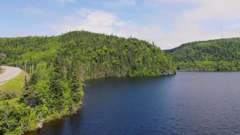 Drone-footage-of-a-lake-as-the-drone-rises-up-high-above-the-lake-with-forest-covered-mountains-in-the-background-and-a-road-with-a-truck-drives-on-the-left