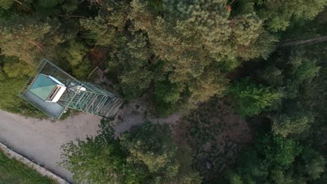 Flyover-old-watchtower-and-dirt-road-surrounded-by-dense-forest,-top-down-aerial-view