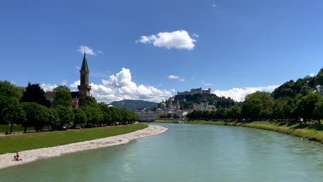 Panning-shot-of-green-Salzach-River-and-Austrian-City-of-Salzburg-in-background-during-sunny-day-with-blue-sky---Rocky-river-shore-and-green-tree-avenue-with-historic-buildings-in-backdrop