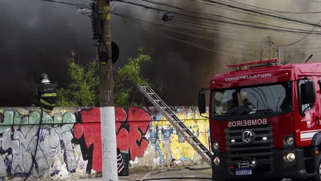 Fireman-suppresses-large-smoky-urban-fire-from-atop-concrete-wall,-on-Tereza-Cristina-avenue,-Ipiranga-district
