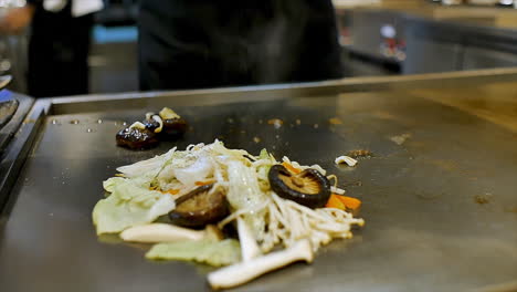 Un-Chef-Japonés-Que-Prepara-Verduras-Mezcladas-Salteadas-En-Una-Estufa-De-Placa-Caliente-Para-Cenar-En-Los-Clientes-En-Un-Restaurante-De-Ambiente