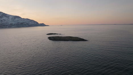 Aerial-drone-orbits-small-rocky-island,-Tromvik-Kvaloya-Northern-Norway-sunset