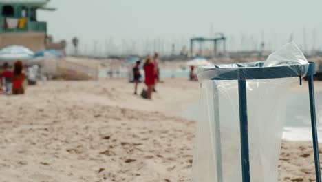 Blauer-öffentlicher-Mülleimer-Mit-Plastiktüte,-Der-Die-Umwelt-Am-Strand-Verschmutzt,-Während-Menschen-Im-Verschwommenen-Hintergrund-Herumhängen
