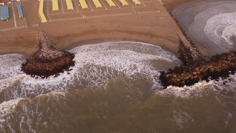 Aerial-top-down-view-of-the-breakwater-at-the-coast,-powerful-water-waves-of-the-Ocean-reaching-sandy-beach-of-Mar-del-Plata-during-sunset,-Argentina
