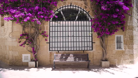 Bench-under-a-window-with-bars-and-climbing-bougainvilleas,Mdina,Malta