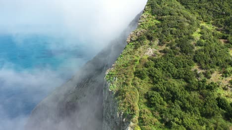 Dünne-Wolken-Ziehen-Langsam-über-Die-Klippe-An-Der-Küste-Der-Insel-Madeira,-Darunter-Türkisfarbenes-Und-Tiefblaues-Wasser-Und-Oben-Grüne,-üppige-Natur,-Auf-Der-Ein-Kleiner-Pfad-Den-Berg-Hinaufführt