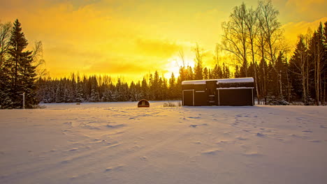 beautiful-yellow-sunset-over-a-winter-coniferous-forest-field,-with-a-cabin-house-and-barrel-sauna