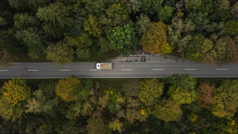 A-fast-driving-lorry-at-a-straight-road-in-a-autumn-colored-forest-street,-filmed-from-above-by-a-drone