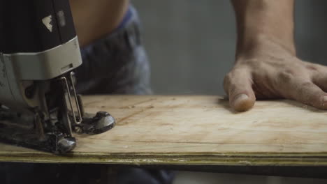 Close-up-shot-of-a-shirtless-worker-cutting-a-wooden-ply-board-with-a-jigsaw-machine-and-making-a-skateboard