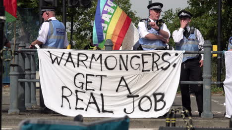 Metropolitan-police-liaison-officers-stand-behind-a-white-banner-with-black-writing-on-that-says,-“Warmongers,-get-a-real-job”-at-a-protest-against-the-DSEi-arms-fair