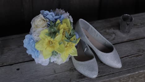 Brides-blue,-white-and-yellow-bouquet-with-her-silver-heels-laid-out-as-a-flat-lay-on-a-wooden-bench