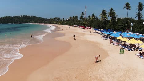 Aerial-dolly-in-of-people-relaxing-in-sand-shore-and-turquoise-sea-surrounded-by-forest-at-daytime-in-Mirissa-beach,-Sri-Lanka