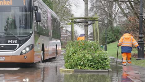 Afroamerikanischer-Busfahrer-Fährt-Auf-Der-überfluteten-Straße-An-Städtischen-Arbeitern-Vorbei,-Die-Nach-Heftigen-Regenfällen-Im-Woluwepark-In-Brüssel,-Belgien,-Den-Abwasserkanal-Reparieren