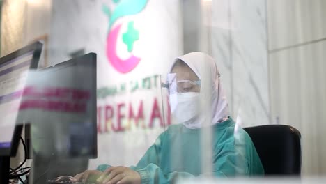 Young-female-worker-wearing-mask-and-hijab-typing-on-computer-at-workplace-in-office-to-enter-patient-data-in-hospital-patient-registration-room